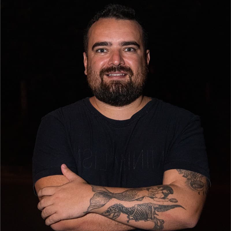 Homem barbudo sorridente de braços cruzados, exibindo tatuagens de sereia e dinossauro nos antebraços.