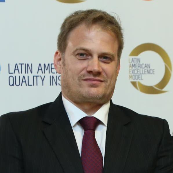 Homem de terno escuro e gravata bordô sorrindo em frente a logotipos institucionais brancos.