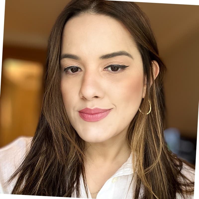 Mulher com cabelos castanhos longos, maquiagem com delineado e batom rosa, usando camisa branca.